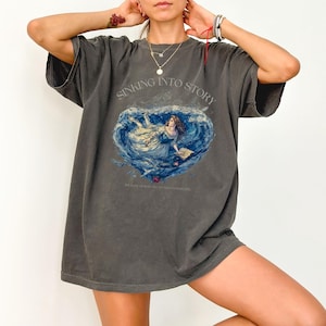 Pode incluir: T-shirt cinza escuro oversized com um gráfico em forma de coração de uma mulher em um oceano azul, com o texto "SINKING INTO STORY" acima e uma linha de texto menor abaixo. A camisa tem mangas curtas e um ajuste folgado.