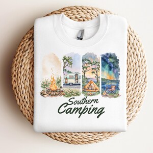 May include: White sweatshirt featuring a watercolor design with camping scenes. The design includes a campfire, a camper, a tent, and a lake scene under a starry sky. The words "Southern Camping" are written in green script.