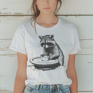 Pode incluir: Camiseta branca com uma ilustração em preto e branco de um guaxinim segurando uma xícara pequena em um prato. O guaxinim é retratado com detalhes de pele e uma expressão focada. A camiseta tem mangas curtas e gola redonda.