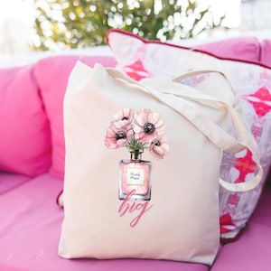 Puede incluir: Bolso tote color crema con un diseño floral. El diseño presenta flores de anémona rosa en un frasco de perfume, con las palabras "Freshly Picked" y "big" impresas en rosa. El bolso tiene asas largas y está sobre una superficie rosa.