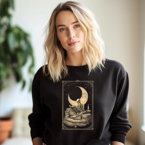 Peut inclure: Sweat-shirt noir à col rond avec un motif détaillé d'un squelette lisant sous un croissant de lune. Le motif comprend des piles de livres et une bougie allumée, le tout dans un cadre décoratif. L'esthétique générale est gothique et mystique.
