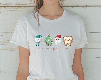 May include: White t-shirt featuring four cartoon dental-themed Christmas characters: an elf tooth, a Christmas tree tooth, a Santa tooth, and a gingerbread tooth. The design is in full color and is centered on the shirt.