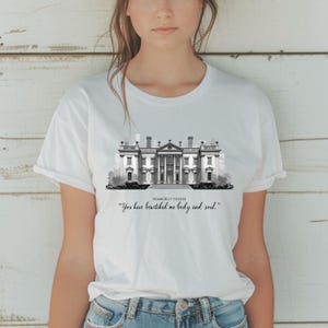 以下が含まれることがあります： 白のTシャツ。黒と白で描かれた壮大な建物のイラスト。テキストは「Pemberly Estate」で、引用句は「You have bewitched me body and soul.」です。