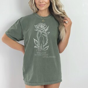 May include: Olive green t-shirt with a white graphic of a rose, a silhouette of a person, and the text "I AM THE VILLAIN IN THIS STORY LITTLE, MOUSE!". The shirt has short sleeves and a crew neck.