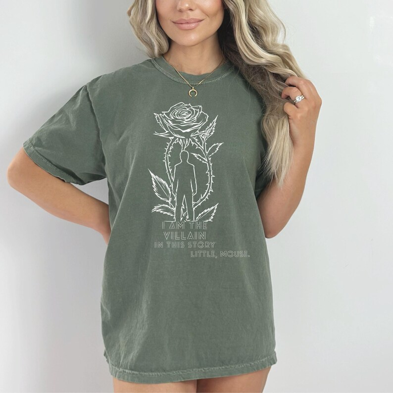 May include: Olive green t-shirt with a white graphic of a rose, a silhouette of a person, and the text "I AM THE VILLAIN IN THIS STORY LITTLE, MOUSE!". The shirt has short sleeves and a crew neck.