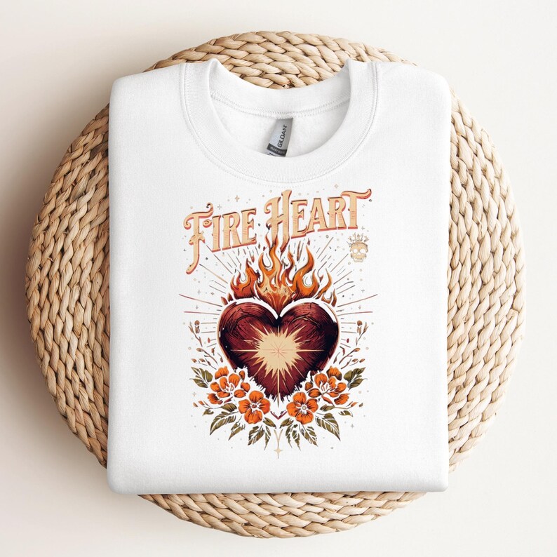 May include: White sweatshirt with a graphic of a heart on fire, and the text "Fire Heart" in an orange vintage font. The design includes orange flowers and leaves. The sweatshirt is folded on a woven, circular surface.