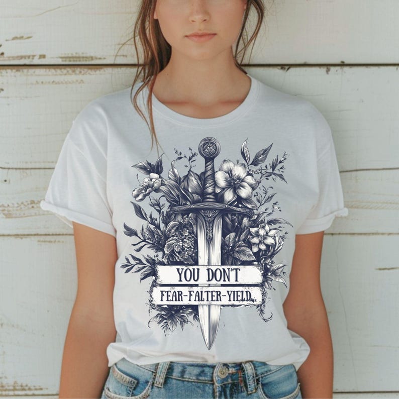 Puede incluir: Camiseta blanca con una ilustraci&oacute;n detallada de una espada rodeada de flores y follaje. El dise&ntilde;o incluye el texto "YOU DON'T FEAR-FALTER-YIELD" en una pancarta. La obra de arte est&aacute; en tonos grises y negros.