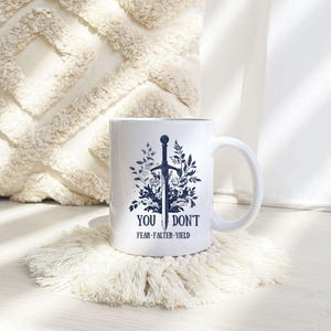 Puede incluir: Taza de cer&aacute;mica blanca con asa, con un dise&ntilde;o de espada azul oscuro rodeado de elementos florales. El texto "YOU DON'T FEAR-FALTER-YIELD" est&aacute; impreso debajo. La taza est&aacute; sobre un posavasos blanco texturizado.