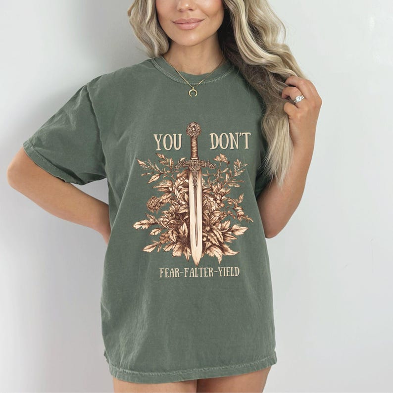 Puede incluir: Camiseta verde oliva con el texto "YOU DON'T" sobre un dise&ntilde;o de espada rodeado de elementos florales. Debajo de la espada, el texto dice "FEAR-FALTER-YIELD". La camiseta tiene mangas cortas y cuello redondo.