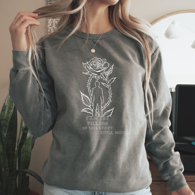 May include: A gray sweatshirt with a white graphic of a rose, a silhouette of a person, and the text "I AM THE VILLAIN IN THIS STORY LITTLE MOUSE". The sweatshirt has long sleeves and a crew neck.