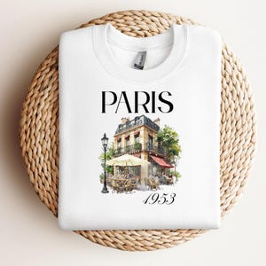 May include: White sweatshirt with a watercolour illustration of a Parisian cafe. The word "PARIS" is above the cafe image, with "1953" below. The cafe has a red awning, outdoor seating, and a street lamp.