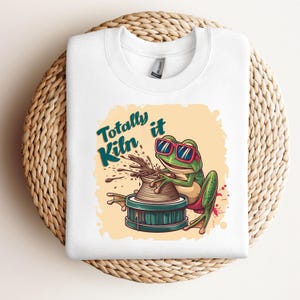 May include: White sweatshirt featuring a cartoon frog wearing sunglasses and creating pottery. The frog is positioned in front of a light orange background with the words "Totally Kiln it" in teal and black.