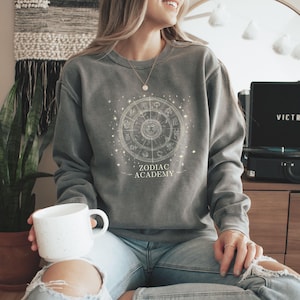 Puede incluir: Una sudadera gris con un diseño de rueda del zodiaco blanca y el texto "ZODIAC ACADEMY". La sudadera se combina con unos vaqueros azules. Se sostiene una taza blanca.