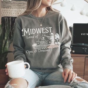 Peut inclure: Un sweat-shirt gris avec le texte "Midwest Land of Opes & Dreams" et un dessin d'une grange, d'un silo et de champs. Le sweat-shirt est porté avec un jean bleu et une tasse blanche.