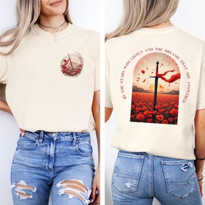 May include: Cream-colored t-shirt with a front graphic of a circle with a mountain and flowers. The back features a sword in a field of red poppies with the text "TO THE STARS WHO LISTEN AND THE DREAMS THAT ARE ANSWERED."
