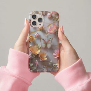 Op de afbeelding: Een telefoonhoesje met een bloemen- en vlinderontwerp. De hoes heeft een marmerachtige achtergrond met roze, gele en perzikbloemen en een vlinder in het midden. De hoes wordt vastgehouden door een persoon die een roze sweatshirt draagt.