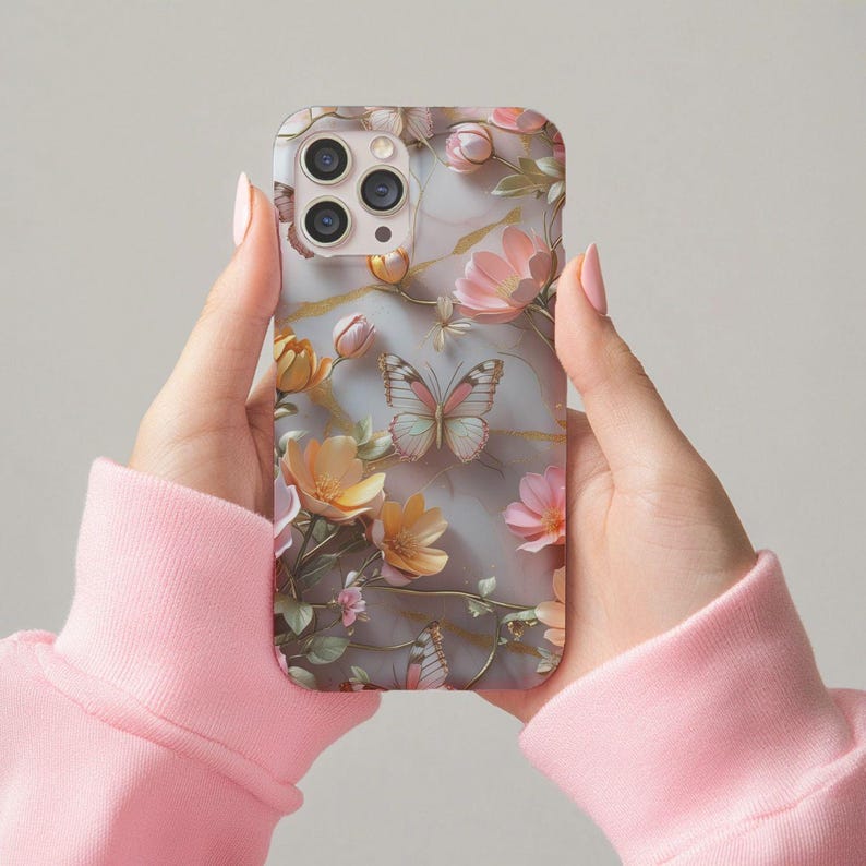 Op de afbeelding: Een telefoonhoesje met een bloemen- en vlinderontwerp. De hoes heeft een marmerachtige achtergrond met roze, gele en perzikbloemen en een vlinder in het midden. De hoes wordt vastgehouden door een persoon die een roze sweatshirt draagt.