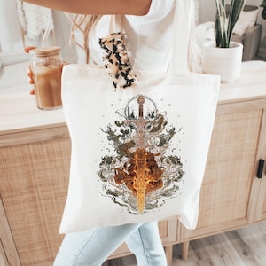Puede incluir: Bolso tote de lona blanca con una ilustración detallada de una espada dorada con diseños intrincados, rodeada de humo arremolinado y elementos similares al fuego. La obra de arte está centrada en la bolsa. La bolsa es sostenida por una persona.