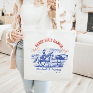 Puede incluir: Una bolsa de tela blanca con un diseño gráfico azul y rojo. El gráfico presenta a una mujer montando un caballo con el texto "Rebel Blue Ranch" en una pancarta azul. El texto "Meadowlark, Wyoming" está debajo del gráfico con "Est. 1800" debajo.