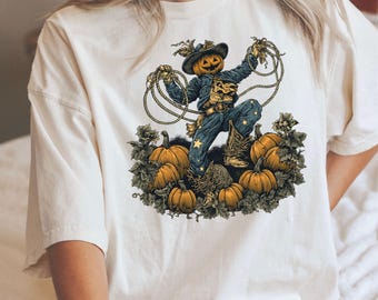 Op de afbeelding: Crèmekleurig T-shirt met een Halloween-thema. Het ontwerp toont een vogelverschrikker met een pompoenhoofd, die een cowboyhoed en -laarzen draagt, omringd door pompoenen. De vogelverschrikker houdt touwen vast, met een sterrenpatroon op de broek.
