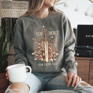 Puede incluir: Una sudadera gris con el texto "YOU DON'T" sobre un dise&ntilde;o de espada rodeado de elementos florales. Debajo de la espada, el texto dice "FEAR-FALTER-YIELD". La sudadera es de color gris apagado.