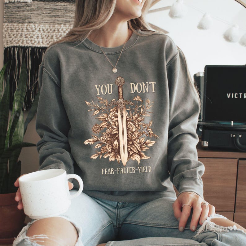 Puede incluir: Una sudadera gris con el texto "YOU DON'T" sobre un dise&ntilde;o de espada rodeado de elementos florales. Debajo de la espada, el texto dice "FEAR-FALTER-YIELD". La sudadera es de color gris apagado.