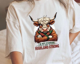 May include: Cream-colored t-shirt featuring a cartoon Highland cow with crossed arms, wearing a red and green plaid scarf. The text reads "I'M NOT STUBBORN, JUST HIGHLAND STRONG" in a bold font.