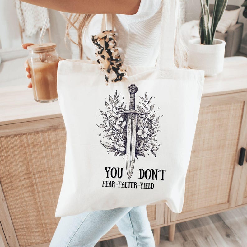 Puede incluir: Bolso tote de color crema con una ilustraci&oacute;n detallada de una espada rodeada de flores y hojas. El texto "YOU DON'T FEAR-FALTER-YIELD" est&aacute; impreso debajo. La bolsa est&aacute; hecha de tela resistente.