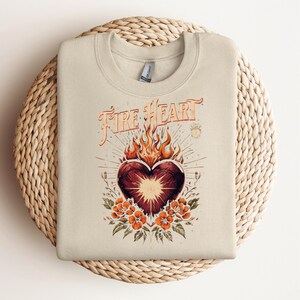 May include: Beige sweatshirt with a graphic design featuring a heart engulfed in flames, surrounded by orange flowers and the text "Fire Heart" in an ornate font. The design has a vintage, bohemian aesthetic.