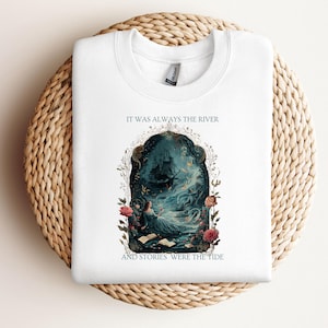 May include: White sweatshirt with the text "IT WAS ALWAYS THE RIVER AND STORIES WERE THE TIDE." The design shows a woman by a river, a ship, and floral elements. The artwork is framed by flowers and foliage, creating a detailed illustration.