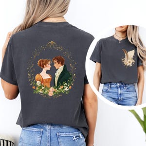 May include: Dark gray t-shirt featuring a romantic illustration of a couple in period clothing, surrounded by a floral wreath. The design is on the back of the shirt. A smaller design of a feather and flowers is on the front.