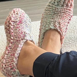 May include: A pair of hand-knitted slippers featuring a chunky, variegated yarn in soft pastel shades of pink, blue, and cream. The slippers have a thick, textured knit pattern with a rounded toe and a low-cut opening, providing a cosy, slip-on design.