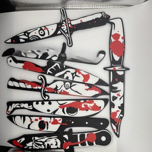 May include: A collection of black and white decorative knives with red blood splatter details. The knives vary in size and design, featuring intricate patterns and shapes. The image showcases a variety of blades, suitable for a themed collection.