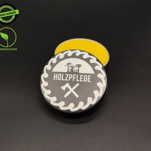 May include: A small, round tin container with a black lid and a white label. The label has a saw blade design with the text "HOLZPFLGE" and a crossed axe symbol. The tin is open and contains a yellow, waxy substance.
