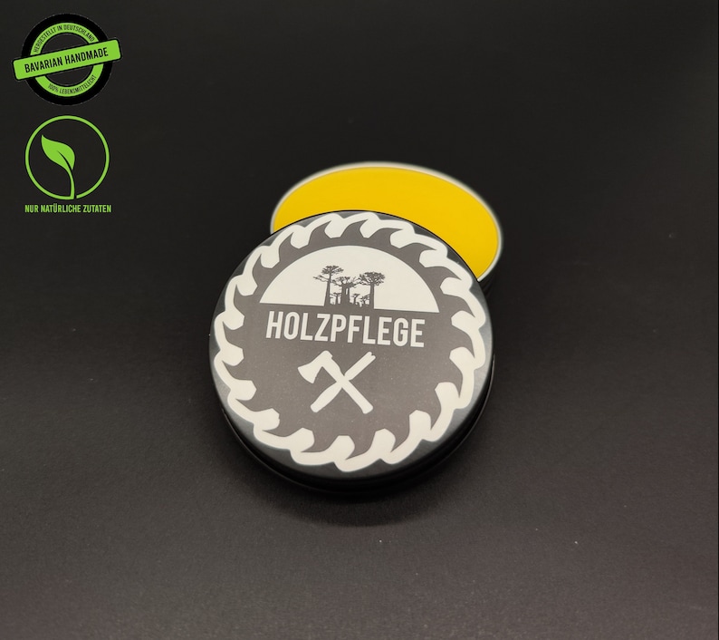 May include: A small, round tin container with a black lid and a white label. The label has a saw blade design with the text "HOLZPFLGE" and a crossed axe symbol. The tin is open and contains a yellow, waxy substance.