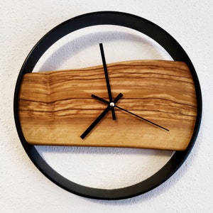 Könnte beinhalten: Eine runde Wanduhr mit einem schwarzen Metallrahmen und einem Zifferblatt aus Naturholz. Die Uhrzeiger sind schwarz, und das Holz hat eine hellbraune Farbe. Die Uhr ist an einer weißen Wand befestigt.