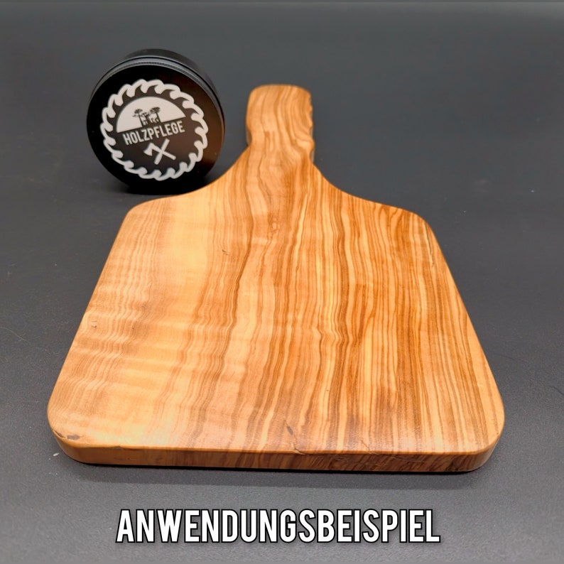 May include: A wooden cutting board with a natural wood grain pattern. The board has a handle and is made of olive wood. The text "ANWENDUNGSBEISPIEL" is printed on the surface below the board.