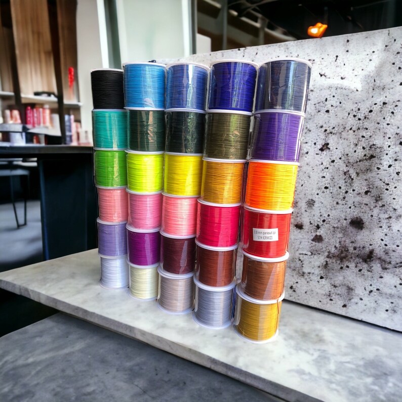 May include: A collection of spools of colorful thread, arranged in a vertical stack. The spools are in various shades of blue, green, yellow, orange, red, pink, purple, brown, and gray. The spools are labeled with the text "1.5 mm paracord 550 (100ft)".