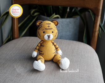 May include: A crocheted tiger doll in yellow and brown, with white paws and muzzle. The doll has black eyes and brown stripes. The text "English Pattern" is in a yellow circle. The doll is sitting on a chair. The text "Nezumi handicraft" is visible.