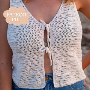 Peut inclure: Haut en crochet crème avec un col en V et une fermeture à nouer. Le devant ouvert est sécurisé par deux liens. Le haut est associé à un jean bleu. Le texte "PATRON PDF" est dans une bulle pêche clair.