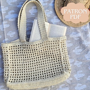 Peut inclure: Un sac fourre-tout en crochet crème à tissage ouvert avec deux poignées, contenant un livre ouvert. Le sac est posé sur une couverture grise à motifs. Un chapeau de paille avec une étiquette "PATRON PDF" est également visible.