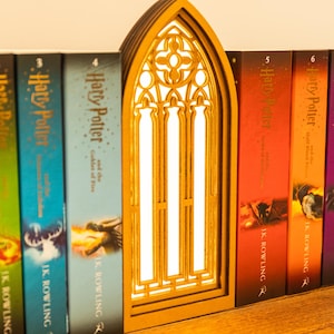 Könnte beinhalten: Ein hölzerner Buchstütze in Form eines gewölbten Fensters mit aufwendigem Design, platziert zwischen Harry Potter Büchern. Die Buchstütze hat einen warmen, goldenen Farbton und ein helles, weißes Inneres, das einen Kontrast zu den Buchrücken bildet.