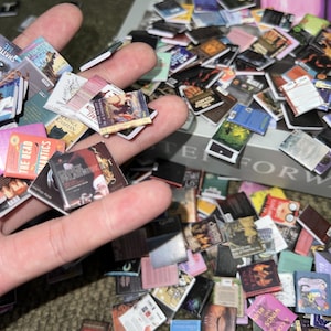 Puede incluir: Una mano sosteniendo una variedad de libros en miniatura, incluyendo t&iacute;tulos como "The Dead", "MANTICS", "Prince Caspian" y "Five Go Off in a Caravan". Los libros est&aacute;n dispuestos en una pila, con algunos de los t&iacute;tulos visibles. El fondo es una caja con la palabra "FORWARD" impresa en ella.