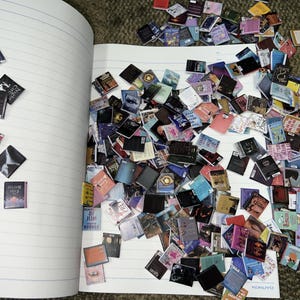 Puede incluir: Un cuaderno con una gran colecci&oacute;n de portadas de libros en miniatura esparcidas por las p&aacute;ginas abiertas. Las portadas son de varios colores y presentan diferentes t&iacute;tulos y autores. El cuaderno tiene p&aacute;ginas rayadas y un fondo blanco.