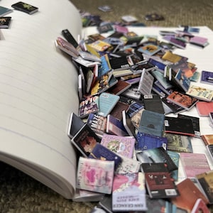 Puede incluir: Un cuaderno con un mont&oacute;n de libros en miniatura que se caen. Los libros son de varios colores y tienen diferentes t&iacute;tulos. El cuaderno est&aacute; abierto y tiene p&aacute;ginas rayadas.