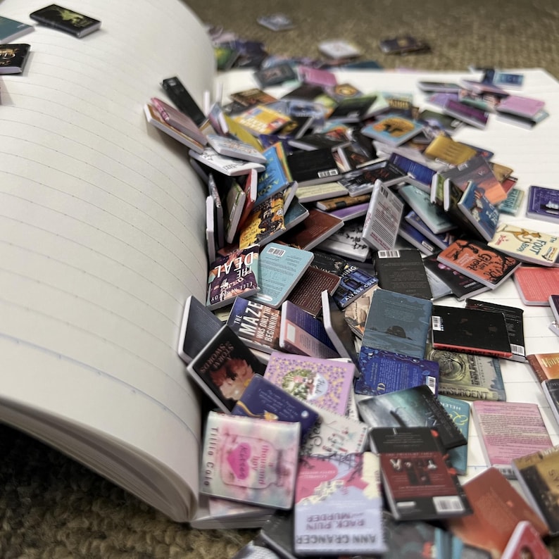 Puede incluir: Un cuaderno con un mont&oacute;n de libros en miniatura que se caen. Los libros son de varios colores y tienen diferentes t&iacute;tulos. El cuaderno est&aacute; abierto y tiene p&aacute;ginas rayadas.