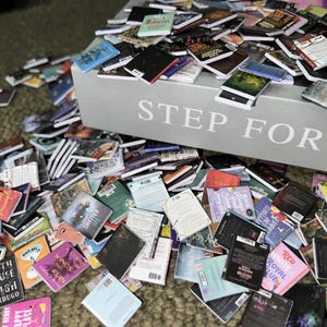 Puede incluir: Una caja grande con las palabras "STEP FOR" impresas en la parte delantera, desbordando de libros de bolsillo en miniatura. Los libros est&aacute;n apilados y esparcidos sobre una superficie marr&oacute;n, con t&iacute;tulos como "Shatter Me", "Wings and Ruin" y "Red, White & Royal Blue" visibles.