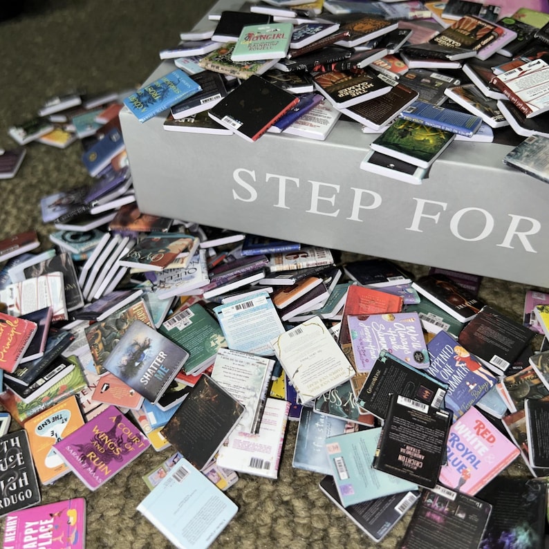 Puede incluir: Una caja grande con las palabras "STEP FOR" impresas en la parte delantera, desbordando de libros de bolsillo en miniatura. Los libros est&aacute;n apilados y esparcidos sobre una superficie marr&oacute;n, con t&iacute;tulos como "Shatter Me", "Wings and Ruin" y "Red, White & Royal Blue" visibles.