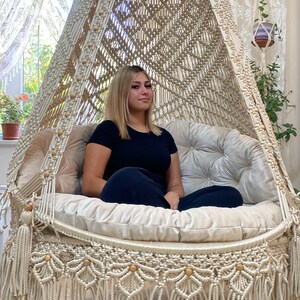 May include: A white macrame hanging chair with a white cushion and tassels. A person is sitting in the chair.