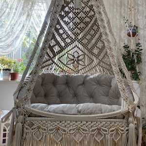 May include: A beige macrame hanging chair with a fringe border and a grey cushion. The chair is suspended from the ceiling and has a decorative tassel.